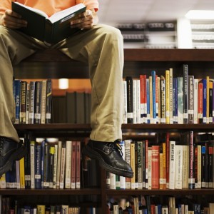 Man Reading Book and Sitting on Bookshelf in Library --- Image by © Royalty-Free/Corbis