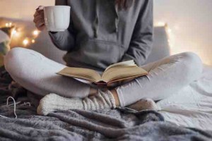 Girl holding cup of hot tea and reading in bed. Around her in bad earphones, book, smart phone. Decorative lights in background.