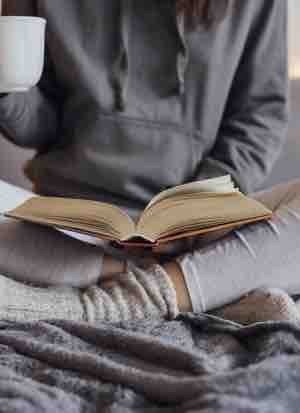 Girl holding cup of hot tea and reading in bed. Around her in bad earphones, book, smart phone. Decorative lights in background.