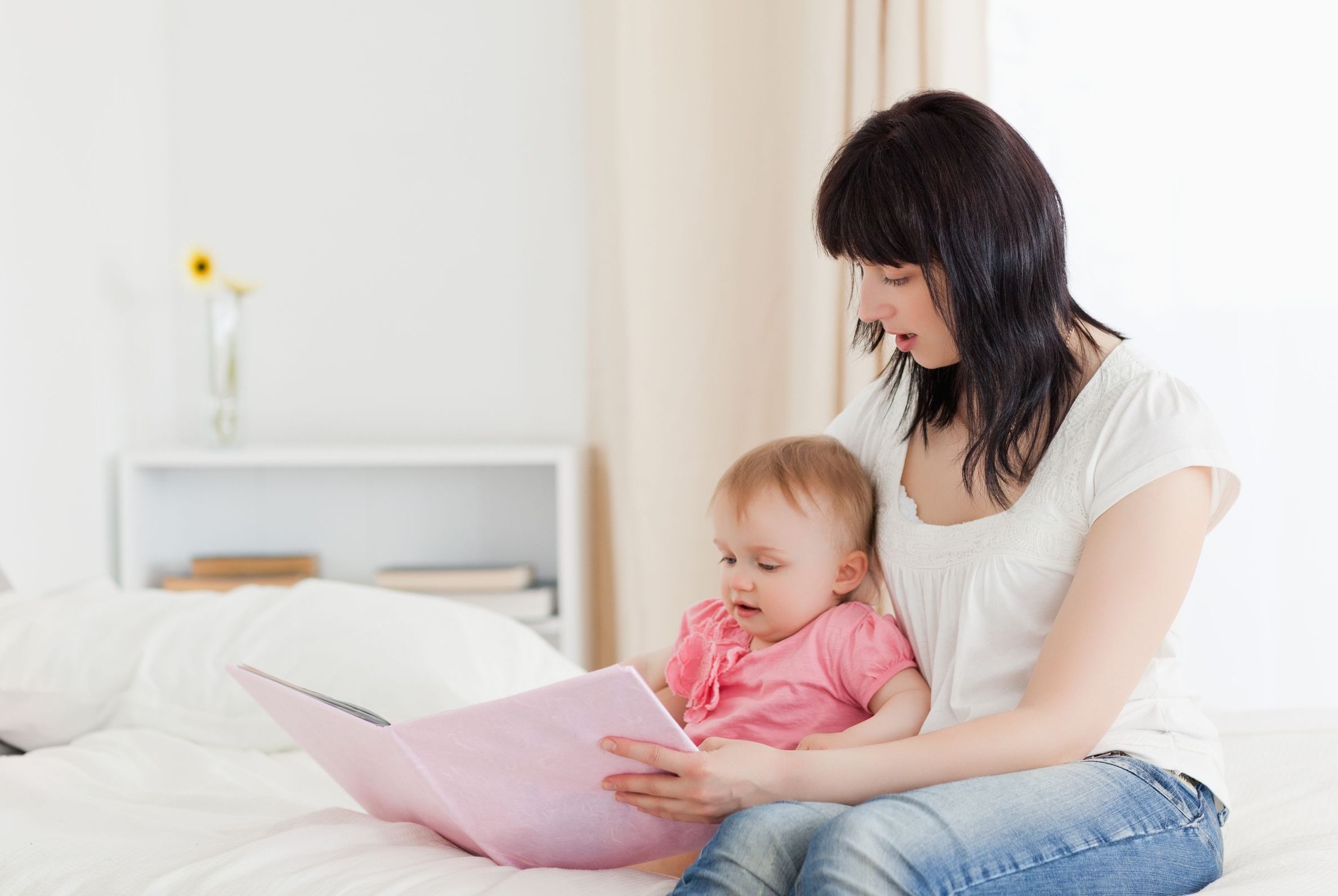 10220941 - attractive brunette woman showing a book to her baby while sitting on a bed in her appartment